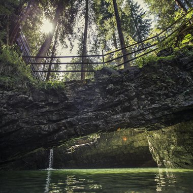 An idyllic place with clear waters and steep cliffs. In the background, tall trees and a sunbeam are visible. | © Kleinwalsertal Tourismus | Oliver Farys