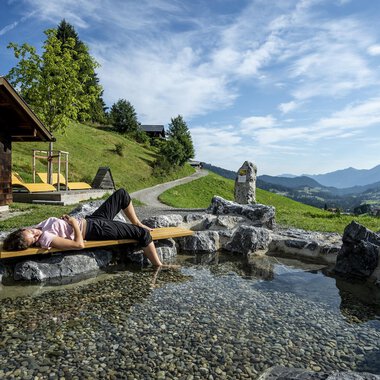 A relaxing scene in the mountains with a person lying by the shore of a small pond. In the background, green hills and a clear sky can be seen. | © Kleinwalsertal Tourismus | Dominik Berchtold