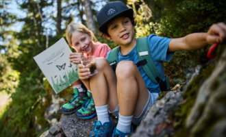 Two children are sitting on a stone in nature. One of the children is holding a book, while the other has a glass with something in their hand.