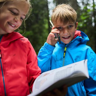 Zwei Kinder stehen im Wald. Das Mädchen lächelt, während der Junge telefoniert und ein Buch hält.