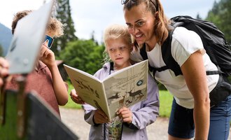 A mother is reading with her children in nature. The children are looking curiously at a book. | © Kleinwalsertal Tourismus | Oliver Farys