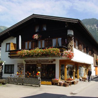 Ein traditionelles Haus in den Bergen, umgeben von Grün. Der Laden im Erdgeschoss bietet handgefertigte Holzschnitzereien an und ist mit Blumen geschmückt. | © Nebelhorn Holzschnitzerei e.K.
