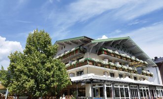 A charming hotel with balconies and colorful plants. In the foreground stands a large tree under a clear blue sky. | © Kleinwalsertal Tourismus | N. Lughammer