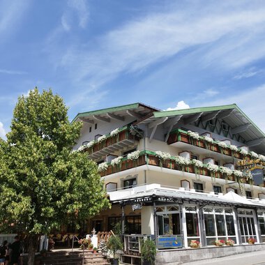 A charming hotel with balconies and colorful plants. In the foreground stands a large tree under a clear blue sky. | © Kleinwalsertal Tourismus | N. Lughammer