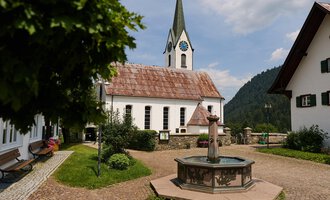 Ein malerischer Dorfplatz mit einem Brunnen im Vordergrund und traditionellen Häusern. Im Hintergrund ist eine Kirche mit einem hohen Turm und einem bewölkten Himmel zu sehen. | © Kleinwalsertal Tourismus | Oliver Farys