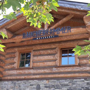 A rustic wooden building with a sign that reads "Mühlenkammer." Surrounded by green leaves and a clear sky. | © Kleinwalsertal Tourismus | Veronika Senn