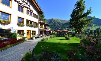 Ein schön gestaltetes Haus mit Balkonen und blühenden Blumen. Im Hintergrund sind grüne Berge und ein klarer Himmel zu sehen. | © Hotel Bellevue | I. Malouvier