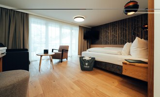 A modern hotel room with wooden flooring and large windows. The furnishings include a bed, an armchair, and a table.