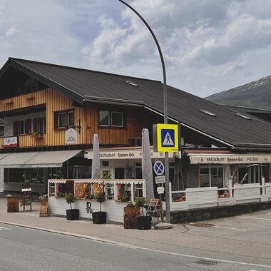 A traditional alpine building with wooden cladding and a restaurant on the ground floor. In the foreground, flower arrangements and traffic signs can be seen. | © Riezlern Eck | Sead Sulejmani