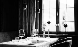 An elegant restaurant table with stylish glasses and a white tablecloth. The windows in the background let in soft light and showcase plant decorations.