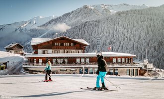 Two skiers are skiing on a snow-covered slope in front of a beautiful mountain hotel. The landscape is surrounded by mountains and Scandinavian forests.
