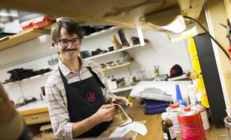 A shoemaker in his workshop smiles as he works on a shoe. In the background, various tools and materials are visible. | © Kleinwalsertal Tourismus | Oliver Farys