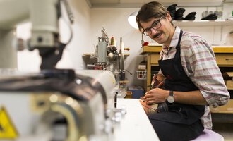 Ein Schuhmacher in seiner Werkstatt, der an einem Paar Schuhe arbeitet. Auf dem Tisch sind Werkzeuge und Maschinen zu sehen. | © Kleinwalsertal Tourismus | Oliver Farys