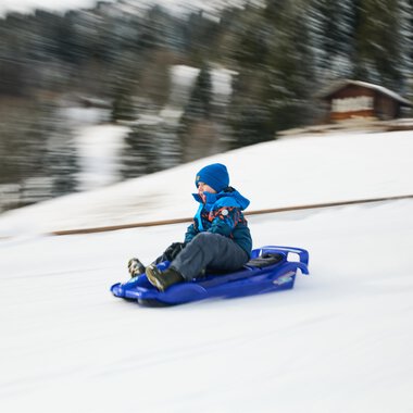 Children are sledding in the snow while adults in warm clothes accompany them. In the background, there is a church and the blue sky is visible. | © Kleinwalsertal Tourismus | Oliver Farys