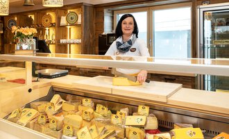 A cheese seller stands behind a counter full of cheese varieties. The shop has a cozy atmosphere with wooden shelves and natural light sources. | © Schönegger Käse-Alm