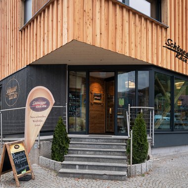 A modern building with a wooden facade and large windows. In front of the entrance stands a sign with an invitation. | © Schönegger Käse-Alm