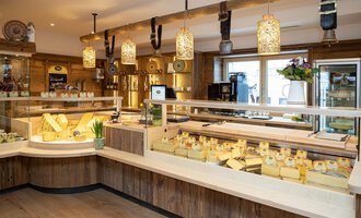 Ein Käsegeschäft mit verschiedenen Käsesorten auf Holzregalen. Die Einrichtung ist rustikal und einladend, mit warmem Licht und dekorativen Elementen. | © Schönegger Käse-Alm