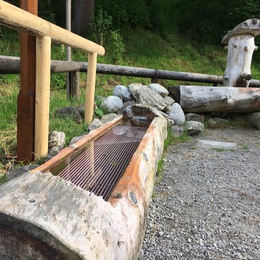 A wooden fountain made of tree trunks stands next to a stone and grass area. Behind it, more wooden structures can be seen, surrounded by trees. | © Kleinwalsertal Tourismus