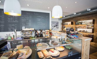 Ein gemütliches Café mit einer Auswahl an Gebäck und Käse. Im Hintergrund ist eine Tafel mit der Speisekarte und Regale mit Getränken zu sehen. | © Kleinwalsertal Tourismus | Frank Drechsel