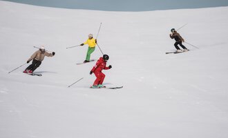 A group of female and male skiers is skiing down the slope. They are wearing various colored ski attire on the snow-covered piste.