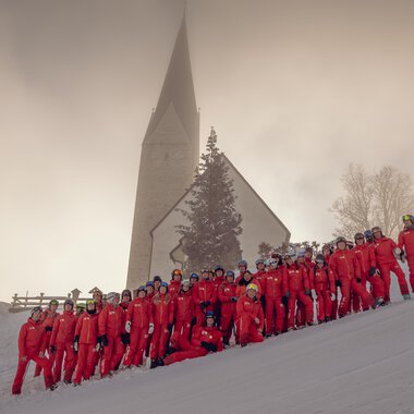 Eine Gruppe von Menschen in roten Skianzügen steht im Schnee vor einer Kirche. Die Landschaft ist winterlich und von Nebel umgeben. | © Skischule Mittelberg | Karl Schuster