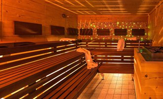 A modern sauna with wooden benches and soft lighting. Relaxation area with multiple seating and decorative light. | © Aparthotel Kleinwalsertal Haller GmbH & Co KG