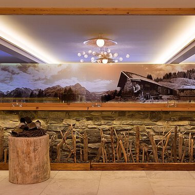 An elegant interior with a stone wall and a large mirror. In the background, there is a large landscape painting featuring a cabin and mountains. | © Aparthotel Kleinwalsertal Haller GmbH & Co KG