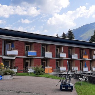 A modern building with multiple floors and balconies. The surroundings are green with a view of the mountains. | © Kleinwalsertal Tourismus | Veronika Senn