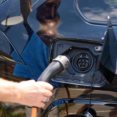 A person is charging an electric vehicle at a charging station. The car is black and the sky is clear. | © Kleinwalsertal Tourismus | Oliver Farys