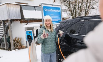 Eine junge Frau lächelt vor einer Ladestation für Elektroautos im Schnee. Sie gibt einen Daumen hoch und trägt eine warme Jacke sowie eine Mütze. | © Kleinwalsertal Tourismus | Oliver Farys