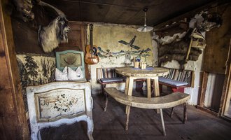 Ein rustikales Zimmer mit einem runden Tisch und Bänken. An den Wänden hängen Tierfelle und dekorative Elemente. | © Kleinwalsertal Tourismus | Oliver Farys