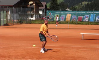 A man is playing tennis on a red court with a view of the mountains. In the background, other players and a tennis wall can be seen. | © Sportverein Kleinwalsertal