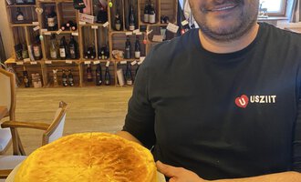 A man is holding a large cake in his hands. In the background, wine bottles are visible on shelves. | © Uusziit | Chantal Kessler