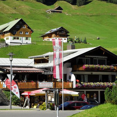 A beautiful alpine building with a terrace and colorful flowers. In the background, green meadows and more traditional houses can be seen. | © Cafe Baad Grund | Roman Schuster