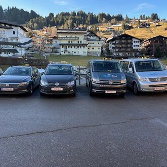 Four taxis are lined up in front of a picturesque mountain landscape. In the background, traditional alpine buildings and wooded hills can be seen. | © Walser Taxi | Josef Düringer