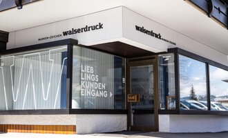 Eine moderne Druckerei mit einem klaren Eingang. Das Schild zeigt den Namen "walserdruck" und die Aufschrift "LIEBLINGS KUNDEN EINGANG". | © Walserdruck KG