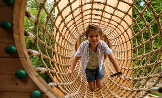A boy is crawling through a large net on a playground. Trees can be seen in the background. | © Kleinwalsertal Tourismus | Oliver Farys