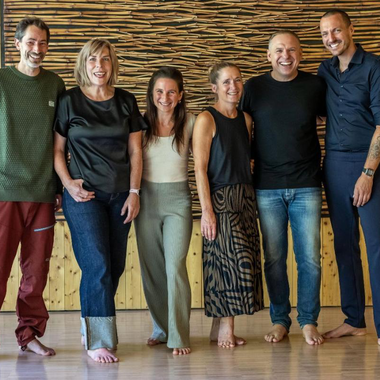 Six people are smiling together in front of a wall clad in wood.  
They are all dressed casually and are standing barefoot on a light floor.