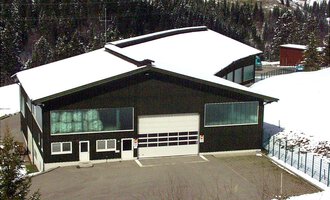 Ein großes, modernes Gebäude mit schwarzer Fassade und schneebedecktem Dach. Umgeben von verschneiten Bäumen und einer ruhigen Landschaft. | © Gemeinde Mittelberg