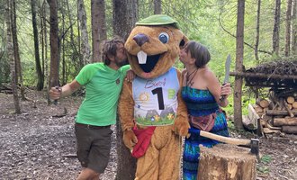 Ein lustiger Maskottchen in Form eines Bibern steht zwischen zwei Personen im Wald. Die Menschen scheinen Spaß zu haben und küssen das Maskottchen auf die Wangen. | © Wildnisschule Kleinwalsertal