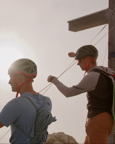 Two climbers securing themselves on a mountain trail with a wooden pole and the sun in the sky. | © Kleinwalsertal Tourismus eGen