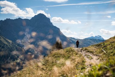 A hiker walking on a trail in the mountains of the Black Forest on a sunny day, surrounded by green grass and mountain peaks in the background. | © Kleinwalsertal Tourismus eGen • Photographer: Andre Tappe