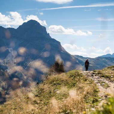 Ein Wanderer geht auf einem Pfad in den Bergen im Schwarzwald bei schönem Wetter, umgeben von grünem Gras und Bergen im Hintergrund. | © Kleinwalsertal Tourismus eGen • Fotograf: Andre Tappe