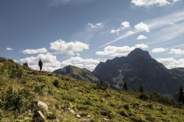 A solitary hiker stands on a lush green hillside overlooking mountains and a blue sky in the Black Water Valley. | © Kleinwalsertal Tourismus eGen • Fotograf: Andre Tappe