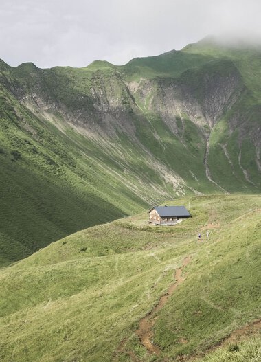 Eine einsame Hütte liegt eingebettet in grüne, sanfte Hügel vor den steilen, bewaldeten Bergkuppen im Hintergrund. | © Kleinwalsertal Tourismus eGen • Fotograf: Andre Tappe