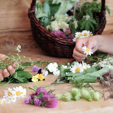 People pick herbs and flowers for a floral or herbal photoshoot at a wooden table, with a basket of flowers in the background. | © Kleinwalsertal Tourismus eGen • Photographer: Christoffer Leitner