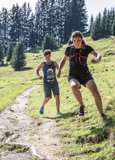 Zwei Trailrunner auf einem Waldweg in den Bergen bei Sonnenschein, einer bei einem Sprung, um Hindernisse zu überwinden. | © Kleinwalsertal Tourismus eGen • Fotograf: Dominik Berchtold