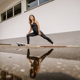 Woman practicing yoga outdoors, reflected in water, in front of a white building. | © Kleinwalsertal Tourismus eGen • Photographer: Dominik Berchtold