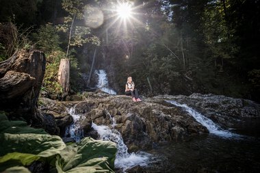 Person sitting on rocks in the Schwarzwasserbach stream in a forest, sunlight shining through trees, water flowing around them. | © Kleinwalsertal Tourismus eGen • Photographer: Dominik Berchtold