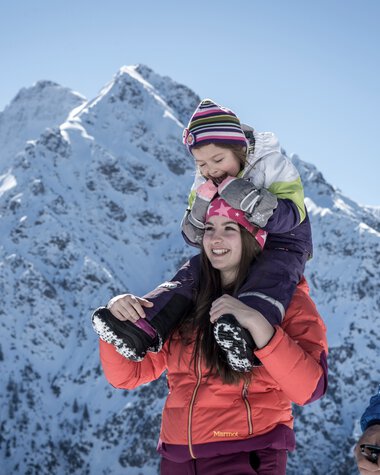 Familie beim Winterwandern in den Bergen, Kinder auf den Schultern der Eltern, mit schneebedeckten Gipfeln im Hintergrund. | © Kleinwalsertal Tourismus eGen • Fotograf: Dominik Berchtold